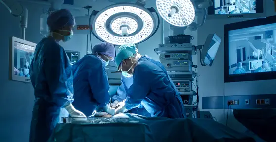 Surgical team performing a procedure in a modern operating room under bright surgical lights with monitoring screens in the background.