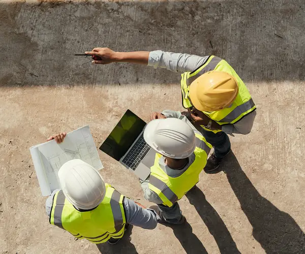 Trabalhadores de capacete e colete refletivo analisando uma planta e um notebook em um canteiro de obras.
