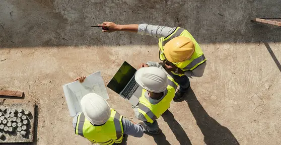 Overhead view of three construction workers in hard hats and safety vests reviewing blueprints and a laptop on a job site.