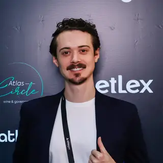 Portrait of a smiling man with dark styled hair, mustache, and goatee wearing a black blazer over a white t-shirt and event credential badge on a black lanyard, making a thumbs-up gesture in front of a blue event backdrop displaying 'Atlas' branding related to wine and governance.