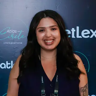 Portrait of a smiling woman with long black hair wearing a dark sleeveless top and event credential badge on a lanyard, posing in front of a blue event backdrop displaying 'Atlas' branding and governance-related text.