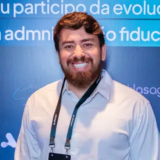 Portrait of a smiling man with reddish-brown hair and a full beard wearing a light blue dress shirt and a black credential badge labeled 'STAFF' on a lanyard, posing in front of a blue event backdrop displaying Portuguese text related to governance and fiduciary administration with Atlas branding.