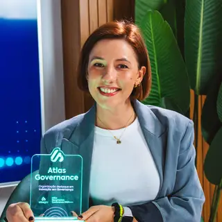 Portrait of a smiling woman with reddish-brown hair wearing a light blue blazer and white ribbed top, holding a modern glass award engraved with 'Atlas Governance' text, posing indoors with green potted plants and a blue LED screen visible in the background.
