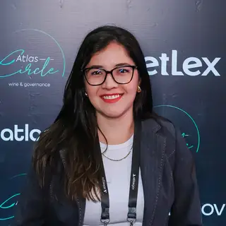 Portrait of a smiling woman with long black hair wearing clear-framed glasses and red lipstick, dressed in a white ribbed top and dark navy blazer with an event credential badge on a lanyard, posing in front of a blue event backdrop displaying 'Atlas' branding and governance-related text.
