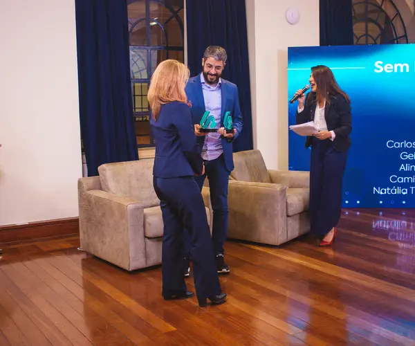 Profissional recebendo troféus de premiação em evento de governança, com tela azul ao fundo exibindo a categoria Sem Fins Lucrativos e o logotipo da Regius, enquanto apresentadora acompanha a cerimônia com microfone