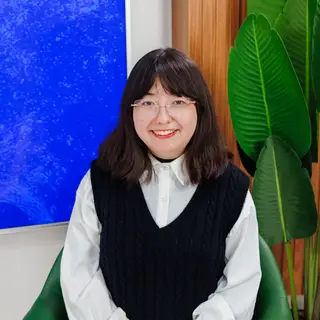 Portrait of a smiling Asian woman wearing glasses, red lipstick, a white long-sleeved shirt, and black sleeveless vest, posing in front of a blue textured wall, wooden door, and green potted plants.