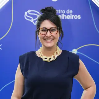 Portrait of a smiling woman with dark hair in a top bun wearing tortoiseshell-framed glasses and a cream-colored chain necklace over a black sleeveless top, posing in front of a blue event backdrop featuring Portuguese text and the IBGC logo with decorative gold curved lines.