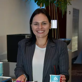 Portrait of a smiling woman with long dark hair wearing a dark pinstriped blazer over a white top and red nail polish, holding an Atlas-branded coffee cup at an indoor event or office setting. Atlas Governance branded materials and cups are visible on the table in front of her, with hanging plants and modern decor visible in the background.