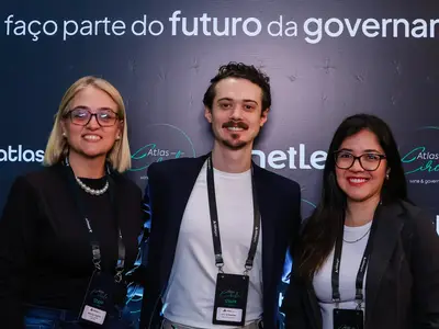 Três membros do time Atlas em pé juntos em um evento de governança, usando crachás de staff, com texto exibindo 'Eu faço parte do futuro da governança' no painel ao fundo