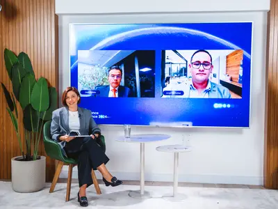 Woman in business attire sitting in a green chair during a virtual meeting displayed on a large blue screen, in a modern office space with wooden paneling and potted plants