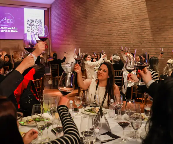 Convidados brindando com taças de vinho durante jantar do evento Atlas Circle, com tela ao fundo exibindo a mensagem "Agora você faz parte do Circle" em ambiente com parede de tijolos
