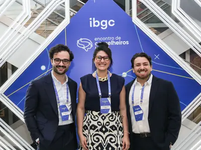 Três profissionais em pé diante de um painel azul do IBGC em um evento de encontro de conselheiros, usando crachás e sorrindo para a câmera