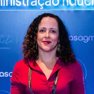 Portrait of a woman with curly reddish-brown hair and red lipstick wearing a bright red blouse and event credential badge on a lanyard, posing in front of a blue event backdrop with Portuguese text related to governance and fiduciary administration.