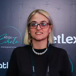 Portrait of a smiling woman with blonde shoulder-length hair wearing black-framed glasses, a pearl beaded necklace, and dangling earrings over a black top with an event credential badge visible, posing in front of a blue event backdrop displaying 'Atlas' branding related to wine and governance.