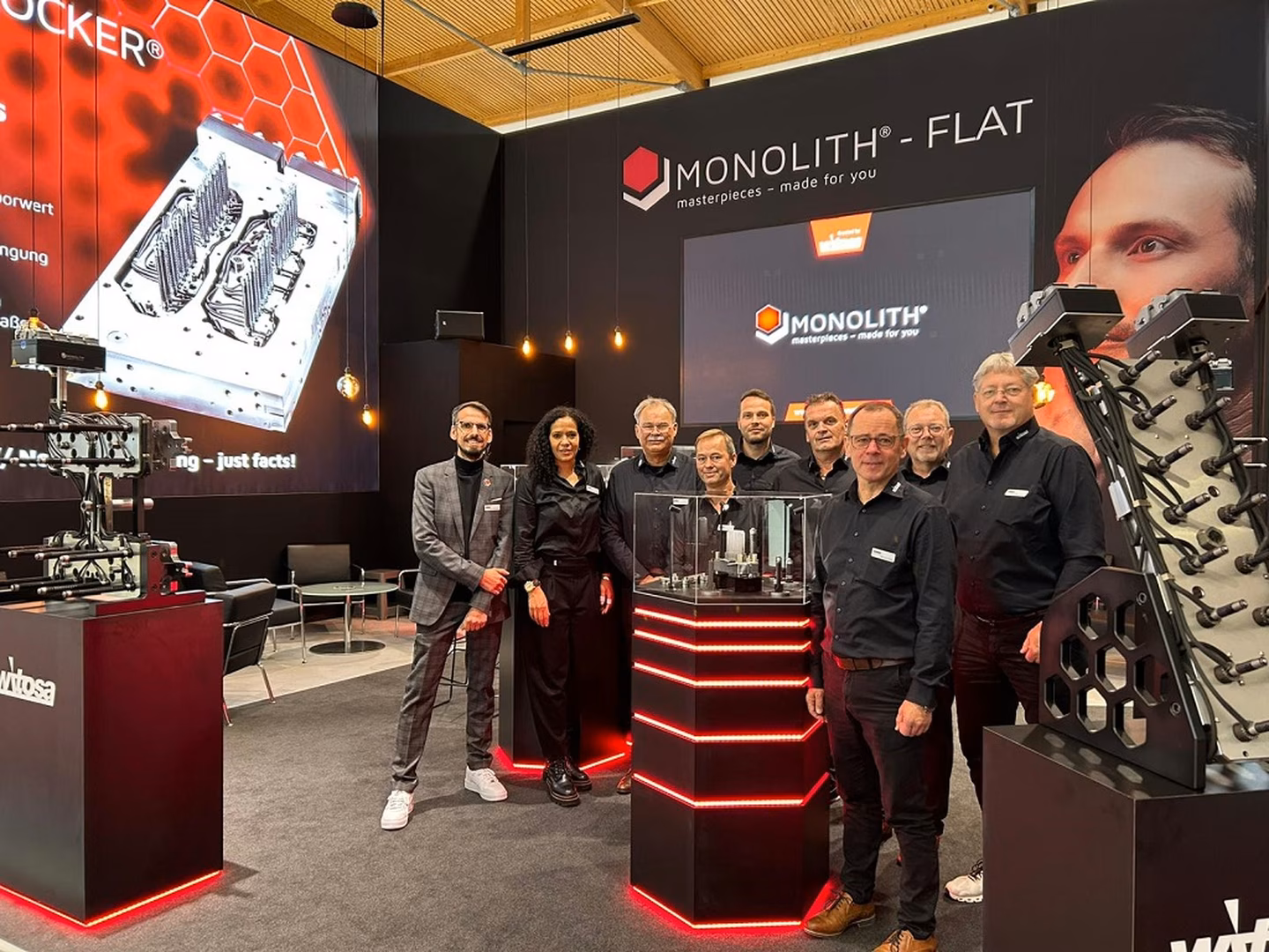 A group of people in black attire stands in a tech exhibition booth, showcasing machinery and Monolith branding, with illuminated displays.