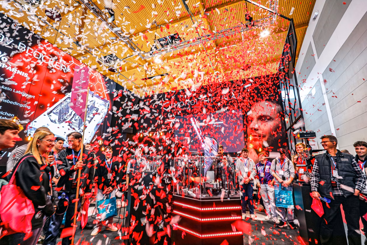 A crowd watches a vibrant display with red and white confetti showering down at a lively tech event. Red LED lights illuminate the stage area.