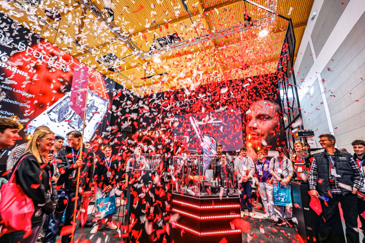 A crowd watches a vibrant display with red and white confetti showering down at a lively tech event. Red LED lights illuminate the stage area.