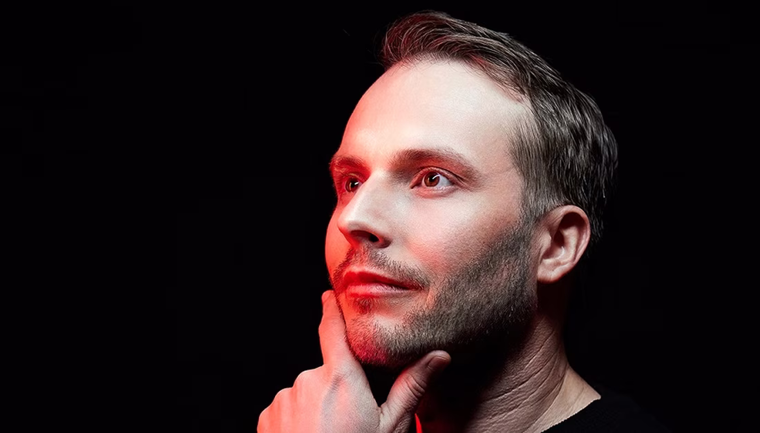 Man with a thoughtful expression, touching his chin, illuminated by red and white light against a black background.