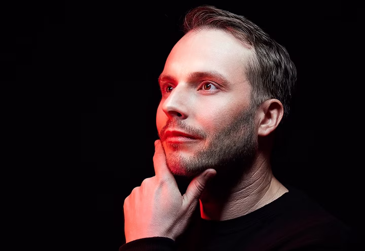 Man with a thoughtful expression, touching his chin, illuminated by red and white light against a black background.