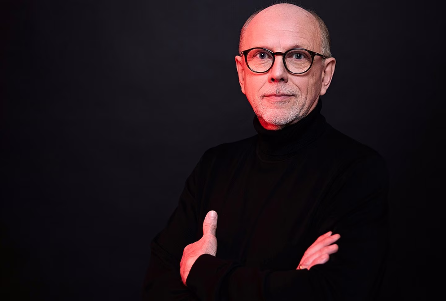 Bald man with glasses and a beard, wearing a black turtleneck, stands confidently with arms crossed against a black background.