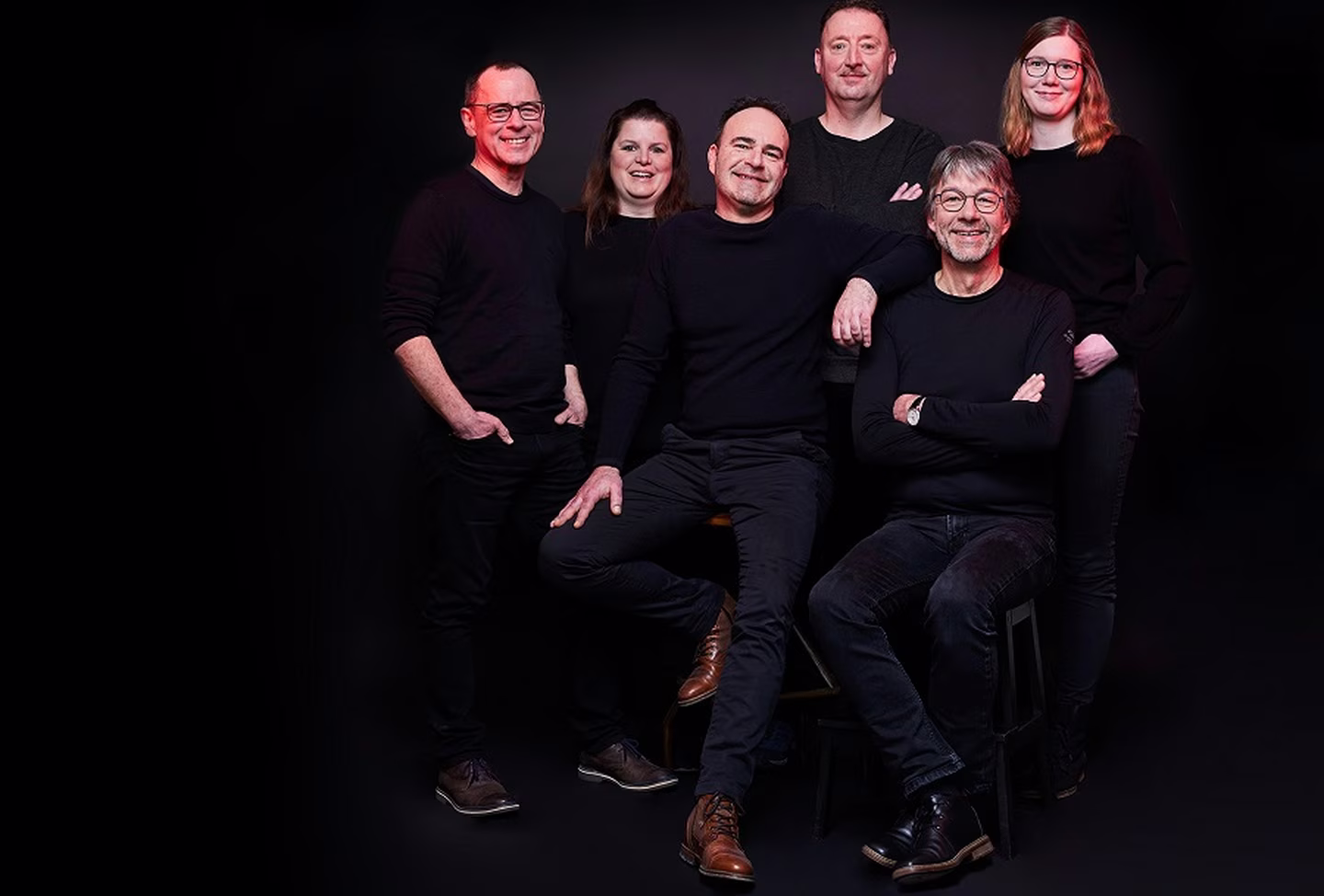 Six people in black clothing, standing and sitting against a dark background, smiling and posing casually.