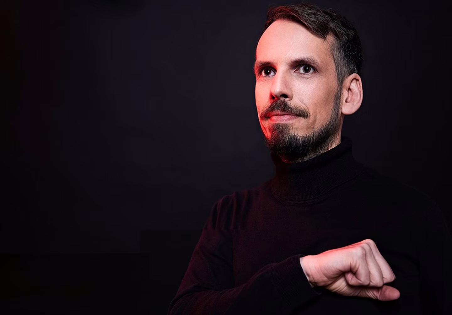 Man with a beard in a black turtleneck gazing upward against a dark background with subtle red lighting on his face.