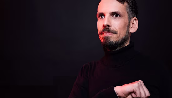 Man with a beard in a black turtleneck gazing upward against a dark background with subtle red lighting on his face.