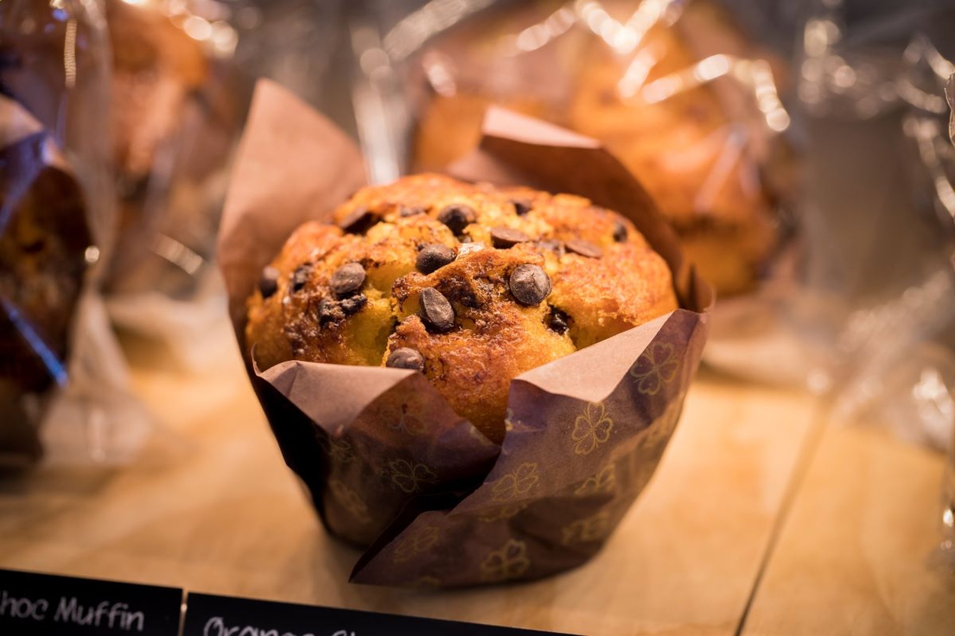 Close-up of a chocolate chip muffin in brown paper wrapping, displayed on a wooden surface with other wrapped baked goods in the background.