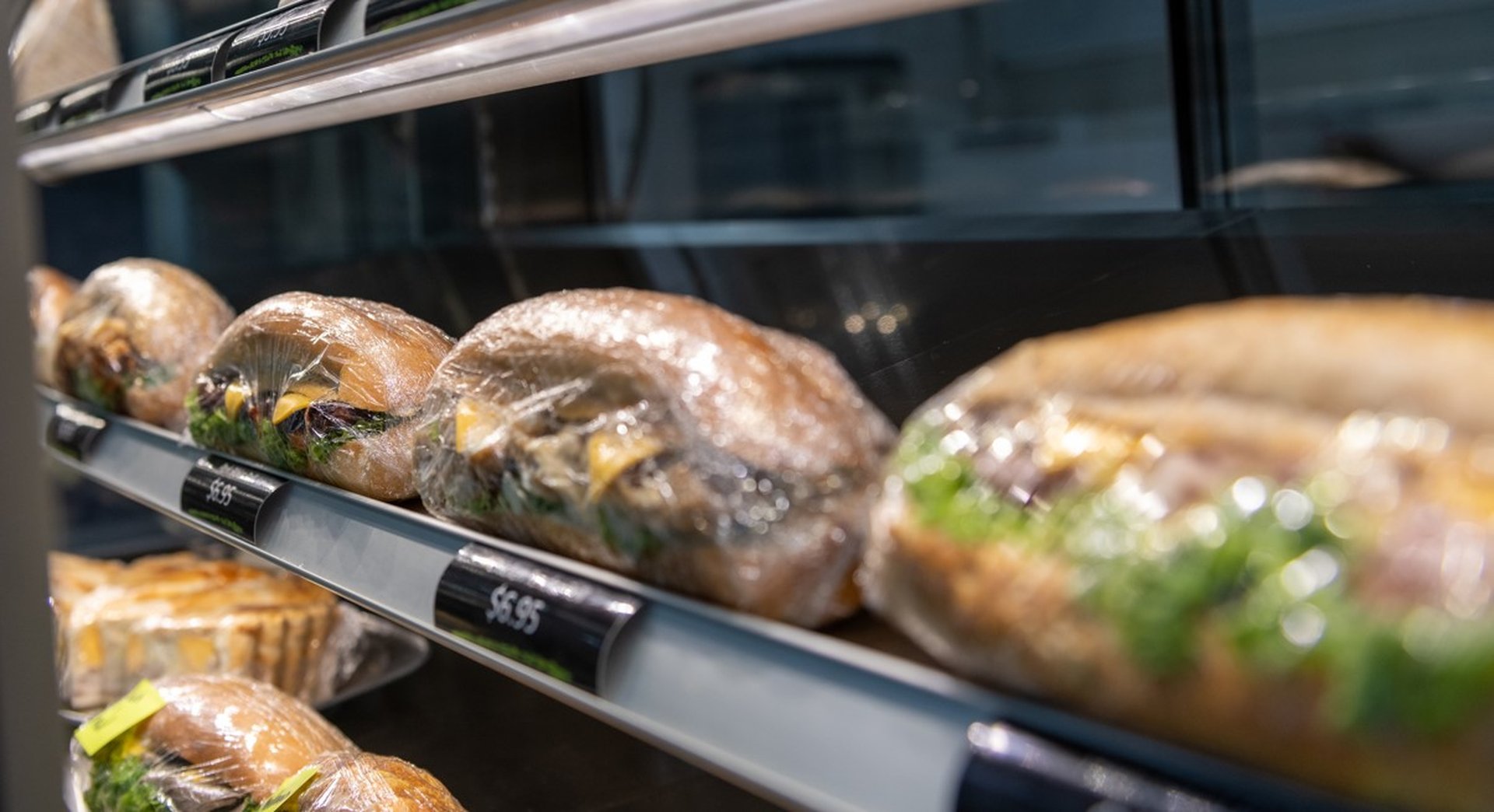 Wrapped sandwiches with various fillings displayed on a shelf in a store, price tags visible, against a blurred background.