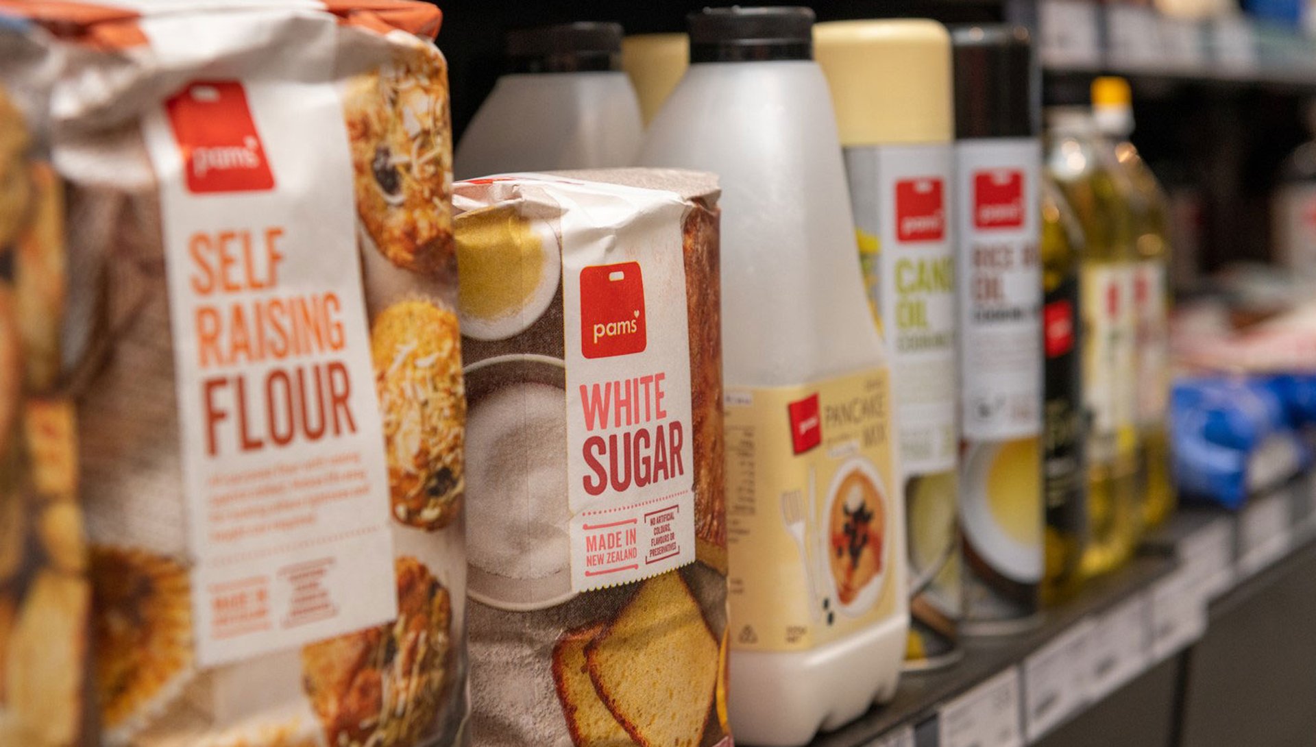 A supermarket shelf displaying various baking goods, including self-raising flour, white sugar, oil, and pancake mix.