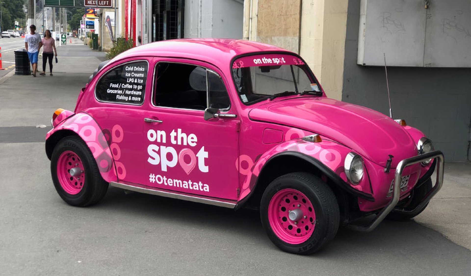 Pink vintage Volkswagen Beetle with "on the spot" branding and hashtag "#Otematata" parked on a city street.