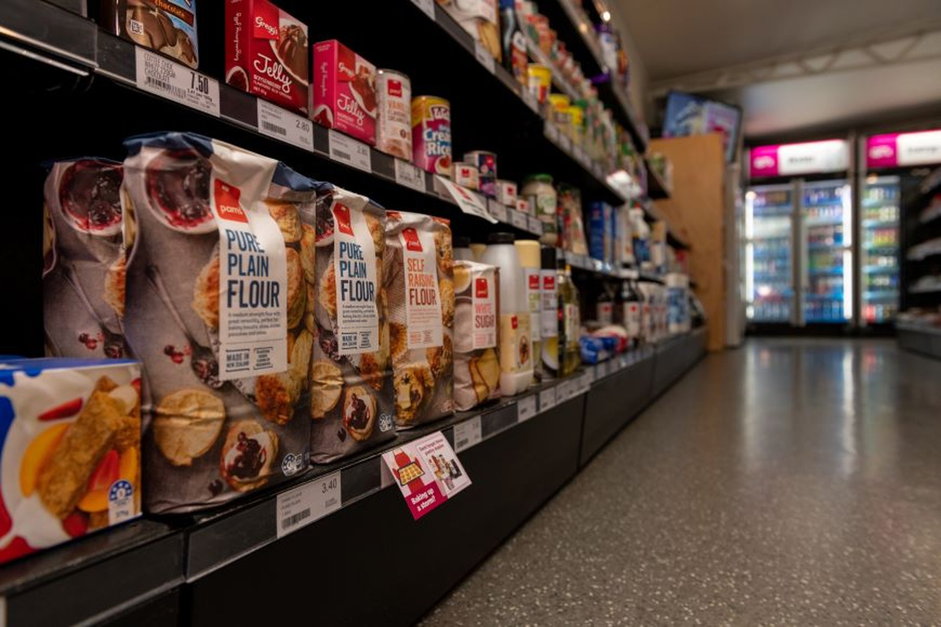A supermarket aisle with various baking goods like flour and sugar on shelves, and refrigerated drinks visible in the background.