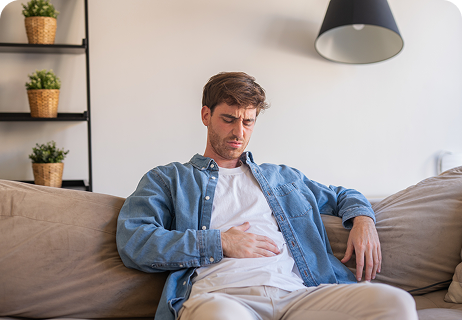A man sitting on a couch, wearing a denim shirt and white pants, holding his stomach with a pained expression.