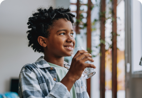 Ein lächelndes Kind mit lockigem Haar in einem karierten Hemd, das ein Glas Wasser in der Hand hält, schaut aus dem Fenster eines sonnendurchfluteten Zimmers.