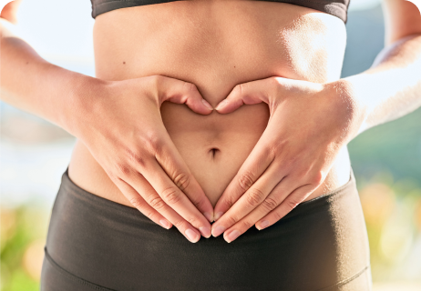 Eine Person, die ein schwarzes Sportoberteil und eine schwarze Sporthose trägt und mit den Händen über ihrem nackten Bauch eine Herzform bildet, im Freien.