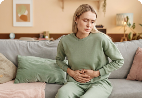 A woman in a green outfit sits on a couch, holding her stomach and appearing to be in discomfort.