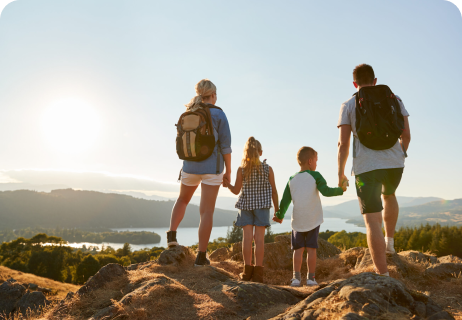 Eine vierköpfige Familie wandert bei Sonnenuntergang auf einem felsigen Hügel und genießt den Blick auf eine malerische Landschaft mit einem See und Bergen im Hintergrund.