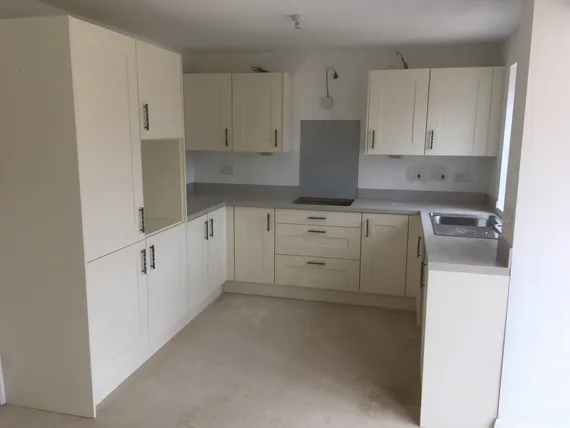 Corner kitchen with white cabinets, grey countertops, built-in stove, and sink. Unfinished floor and walls painted white. Minimalist design.