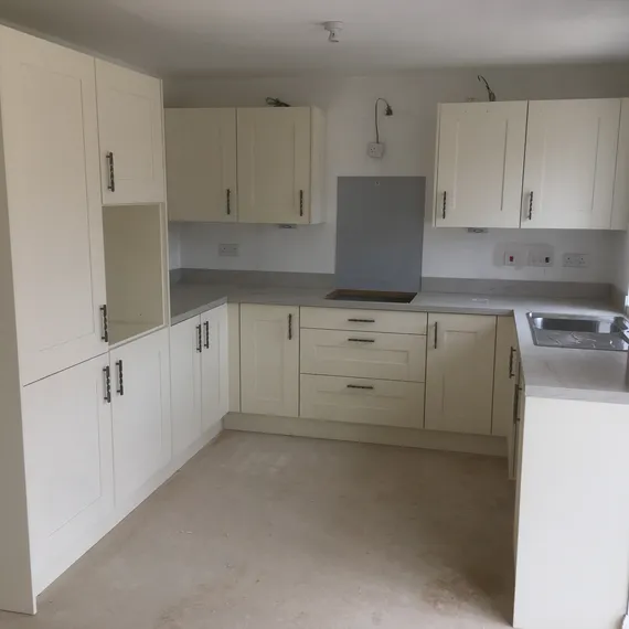 Corner kitchen with white cabinets, grey countertops, built-in stove, and sink. Unfinished floor and walls painted white. Minimalist design.