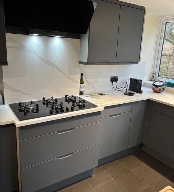 Modern kitchen with gray cabinets, a black stovetop, marble backsplash, wine bottle, and a window showing trees.