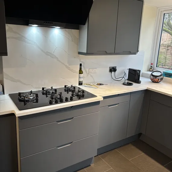Modern kitchen with gray cabinets, a black stovetop, marble backsplash, wine bottle, and a window showing trees.