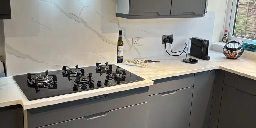 Modern kitchen with gray cabinets, a black stovetop, marble backsplash, wine bottle, and a window showing trees.