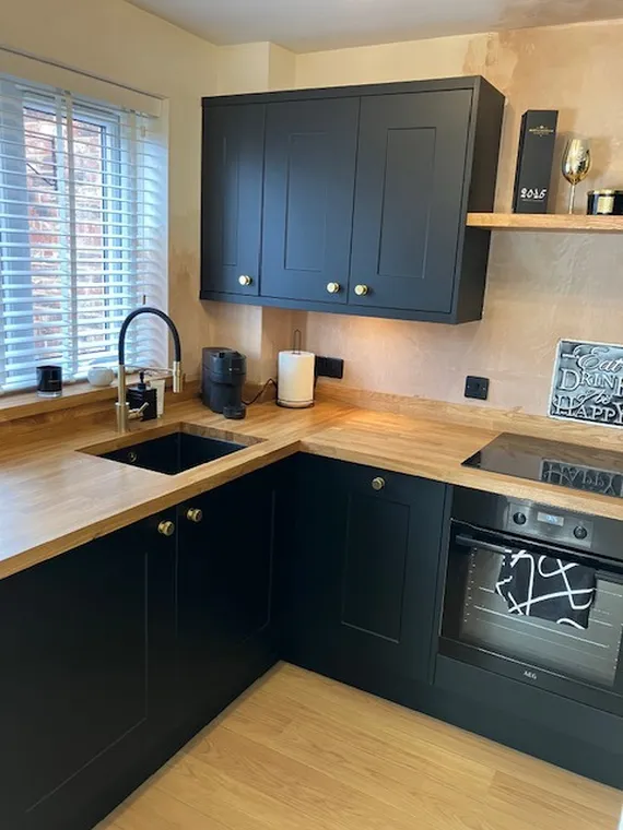 Modern kitchen with black cabinets, wooden countertops, a sink, stovetop, oven, and window with blinds. Warm lighting and minimal decor.