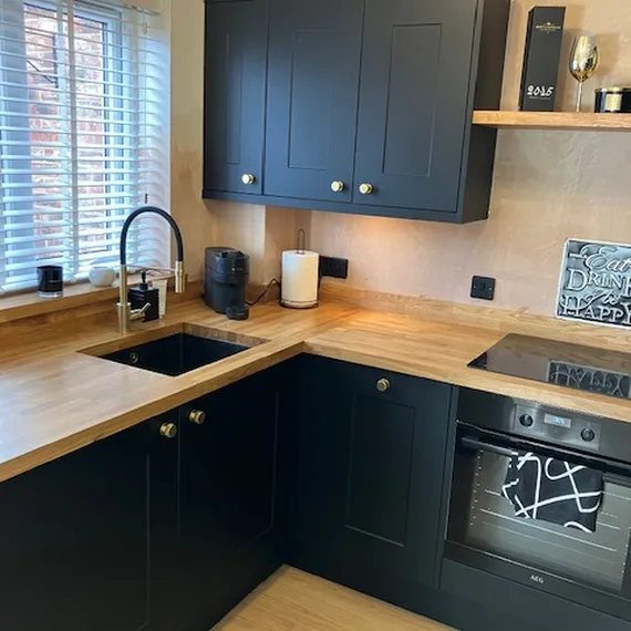 Modern kitchen with black cabinets, wooden countertops, a sink, stovetop, oven, and window with blinds. Warm lighting and minimal decor.