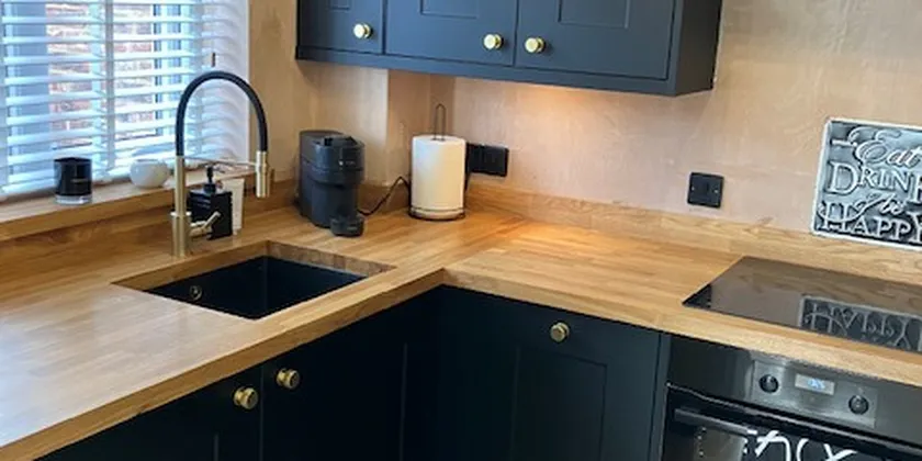 Modern kitchen with black cabinets, wooden countertops, a sink, stovetop, oven, and window with blinds. Warm lighting and minimal decor.