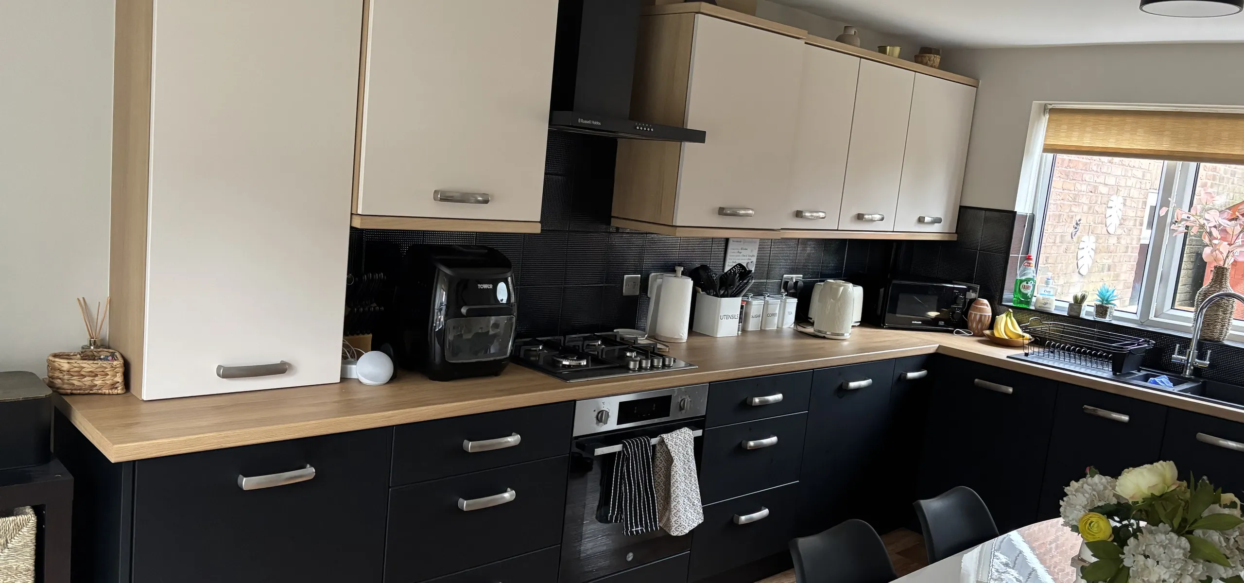 Modern kitchen with wooden and black cabinets, stove, coffee maker, and sink. Sunlight illuminates the counters and dining table.