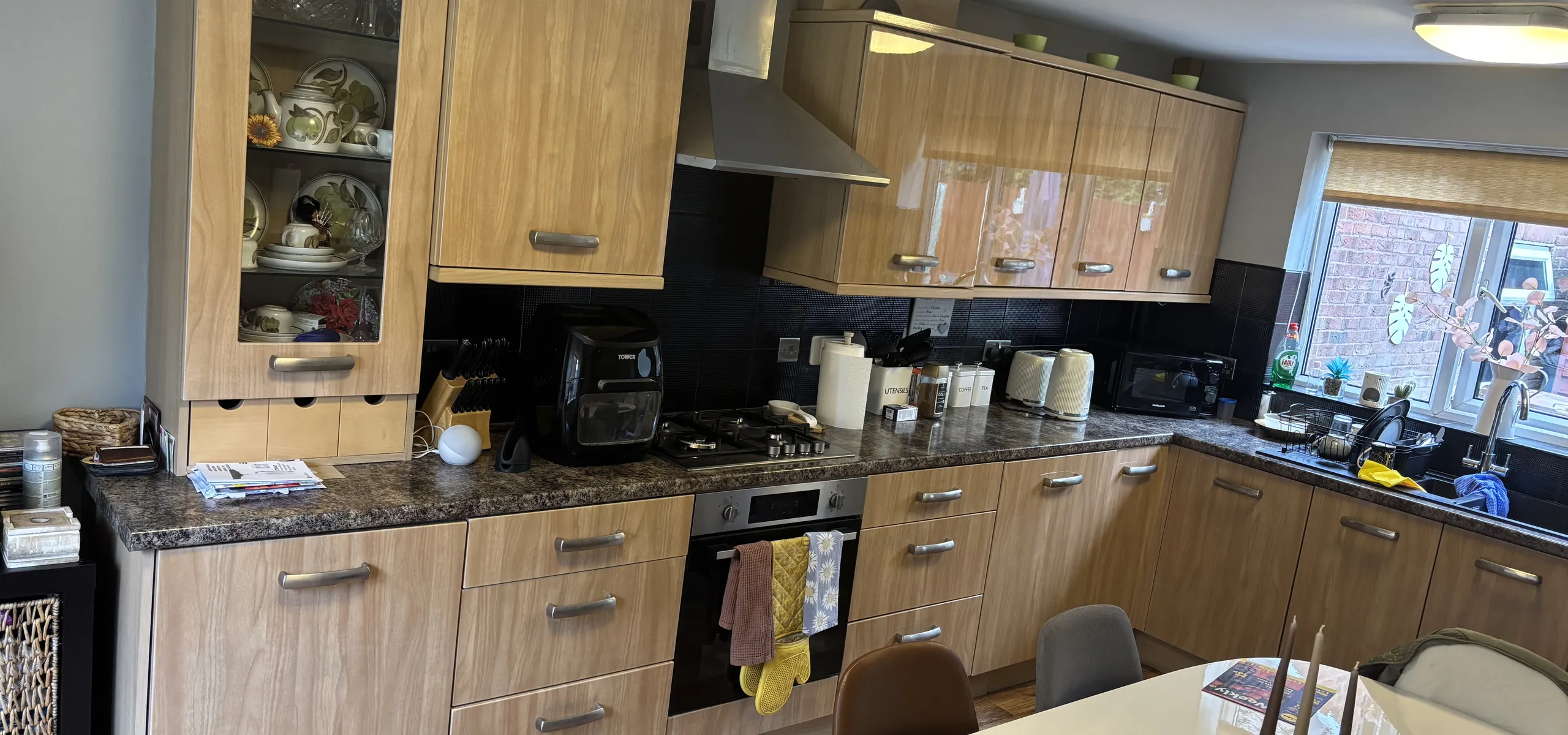 Outdated kitchen with wooden cabinets, black countertops, and appliances. A dining table with papers and magazines is in the foreground.