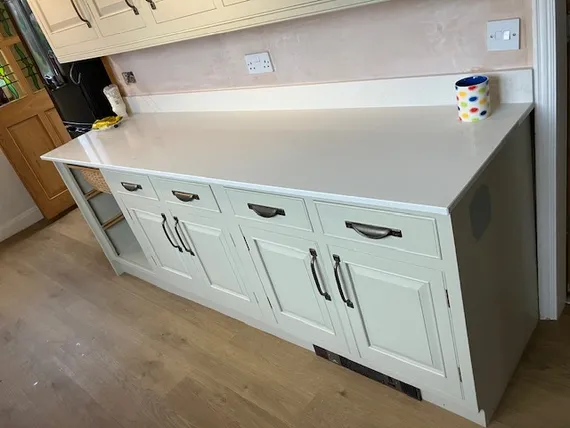 A kitchen counter with a white top, cream cabinets, drawers with dark handles, and a colorful mug beside an electrical outlet.