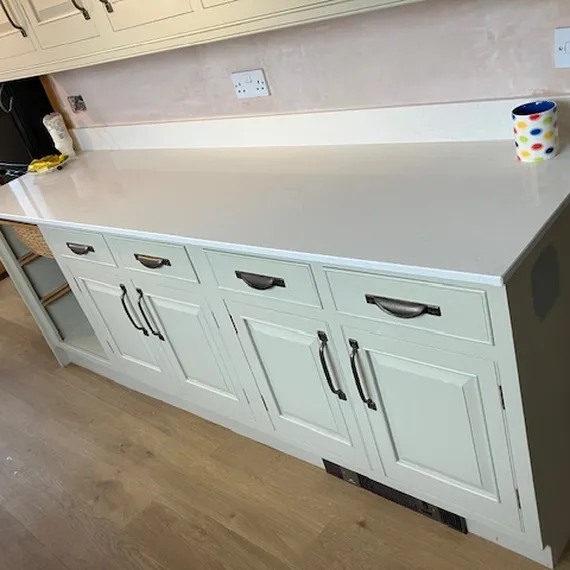 A kitchen counter with a white top, cream cabinets, drawers with dark handles, and a colorful mug beside an electrical outlet.