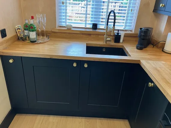 Modern kitchen corner with black cabinets, wooden countertop, sink with faucet, liquor bottles, glasses, coffee maker, and window with blinds.